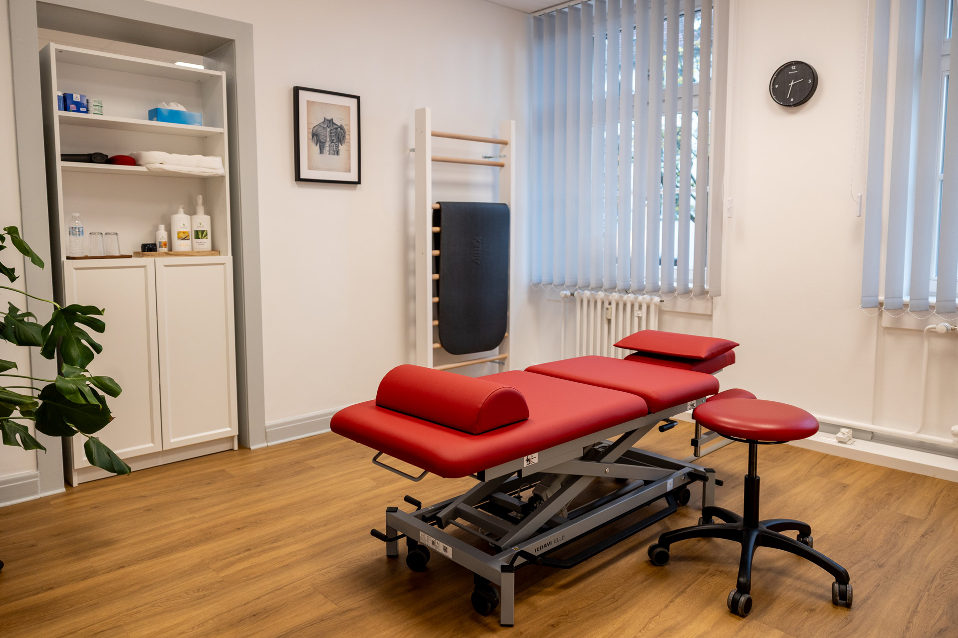 Ein heller Therapieraum mit roter Liege, Holzboden und vielen Fenstern